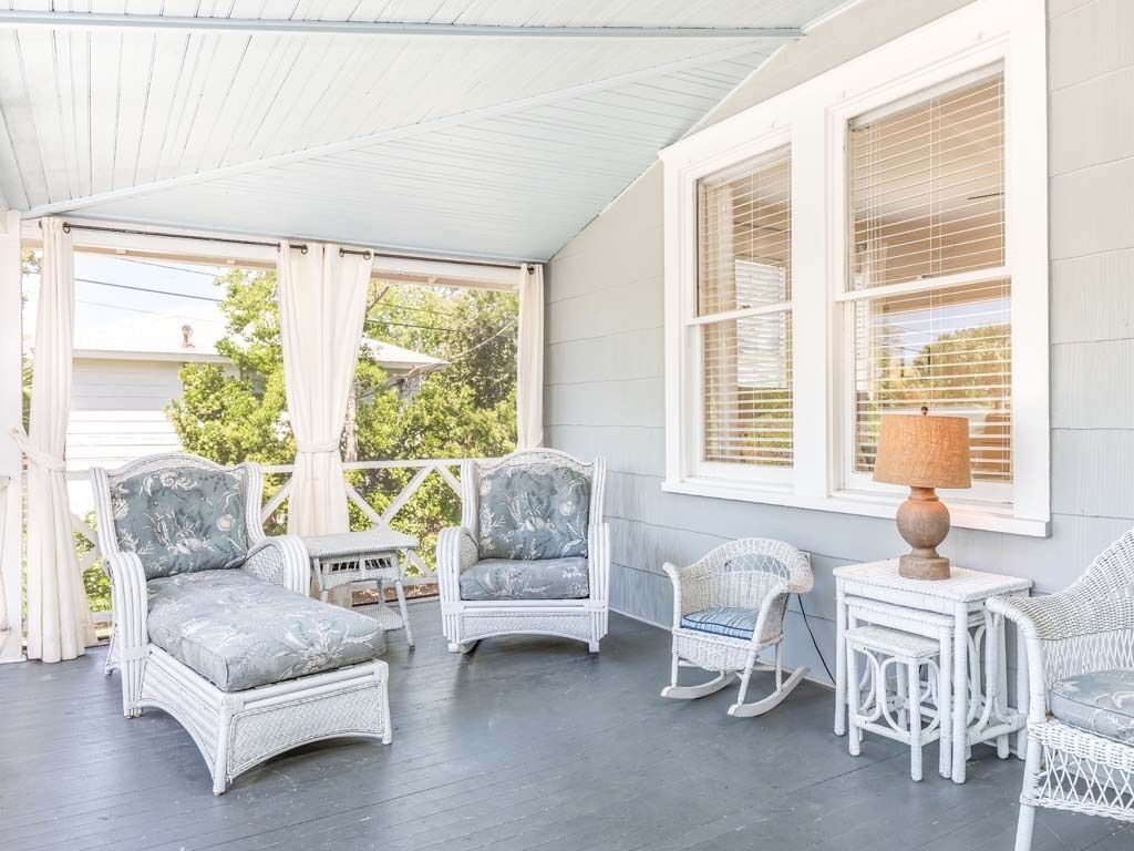 A covered porch with wicker furniture, including chairs and a rocking chair, and a small table with a lamp.