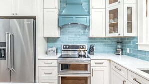 A modern kitchen with white cabinets, a stainless steel refrigerator, a built-in oven, and a blue-tiled backsplash. - Butler Beach Cottage by Tybee Vacation Rentals (Tybee Island)