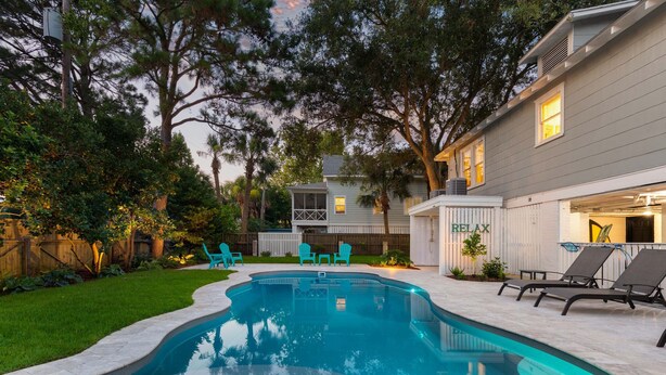 A swimming pool with a curved edge, surrounded by a grassy lawn and trees. There are two outdoor chairs and a lounge chair near the pool. - Butler Beach Cottage by Tybee Vacation Rentals (Tybee Island)