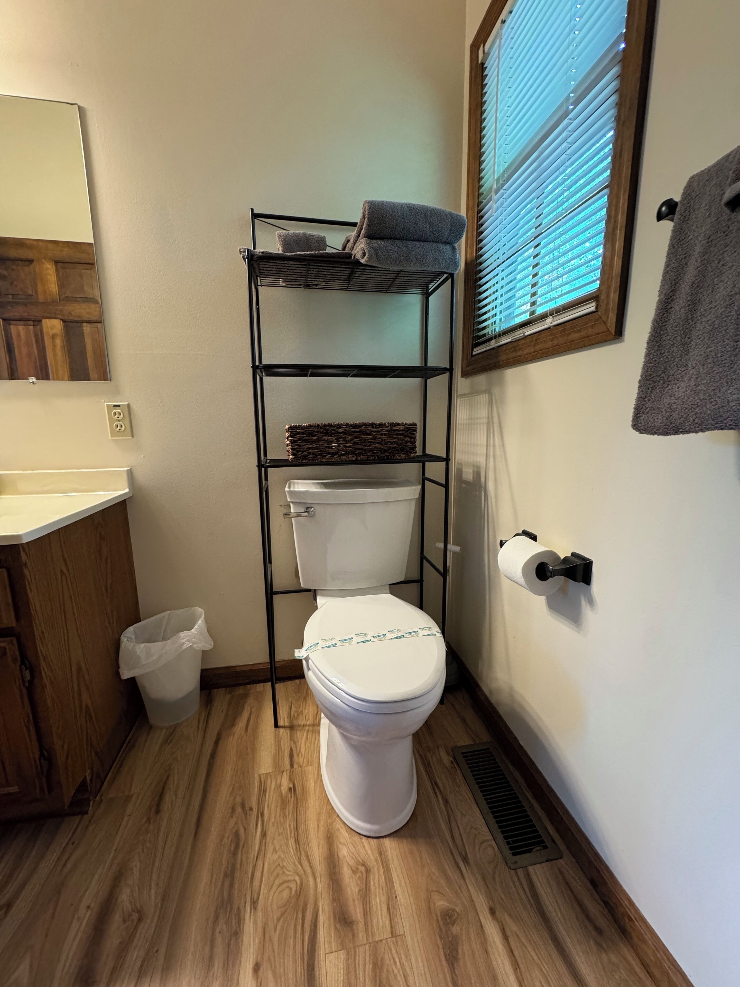 A bathroom with a toilet, a shelf unit with towels, a trash bin, and a window with blinds.