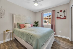 Ein Schlafzimmer mit Bett, Nachttisch, Wanddeko aus Makramee, Deckenventilator, Fenster mit Vorhängen und einem Sonnenuntergangsbild.