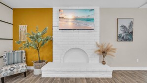 Un salon avec une cheminée en briques blanches, un téléviseur à écran plat fixé au-dessus, un fauteuil à motifs, une plante en pot et une décoration murale en macramé.