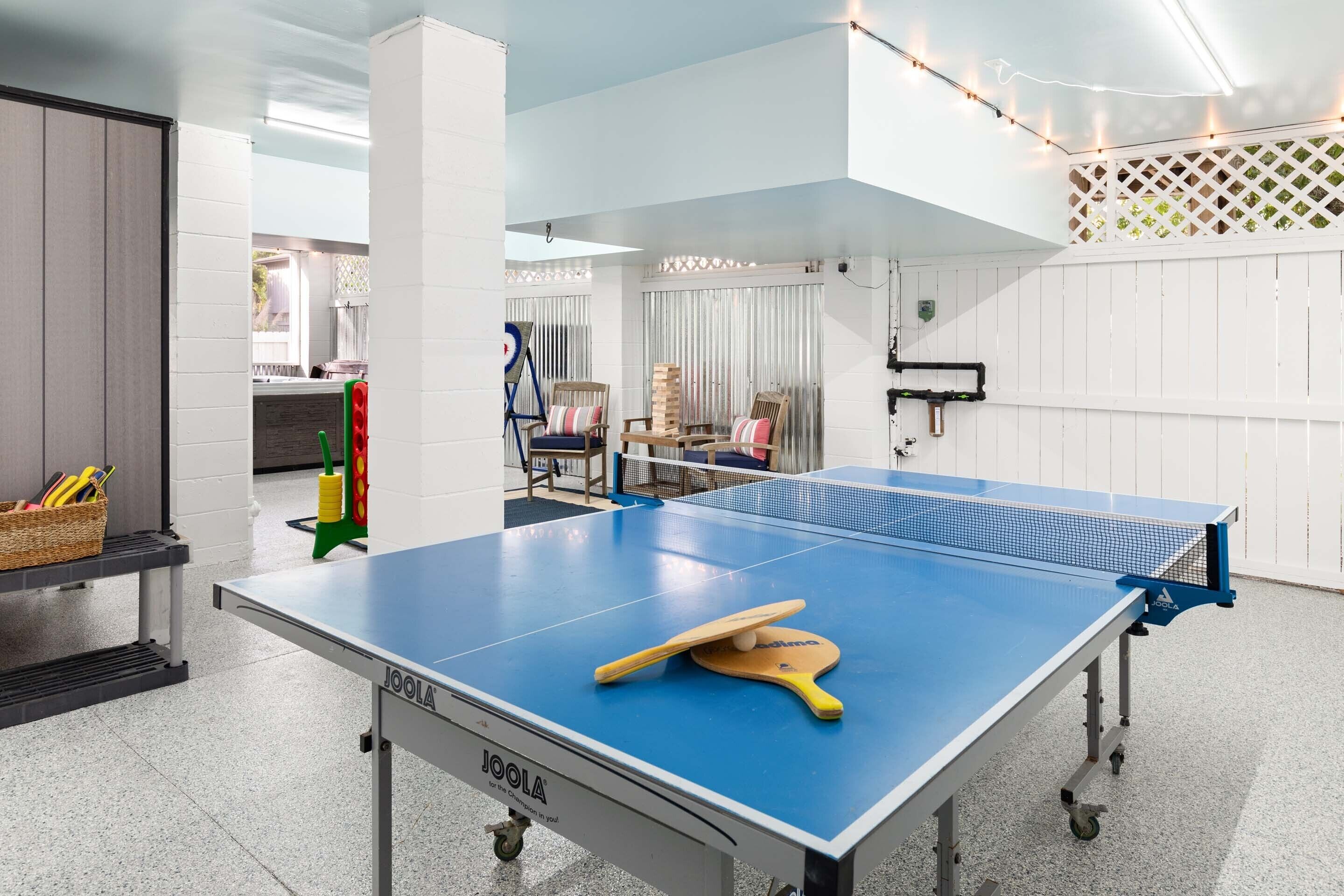A ping pong table with a paddle and ball, a basket of sports equipment, and a table tennis net in a room with white walls and a ceiling light fixture.
