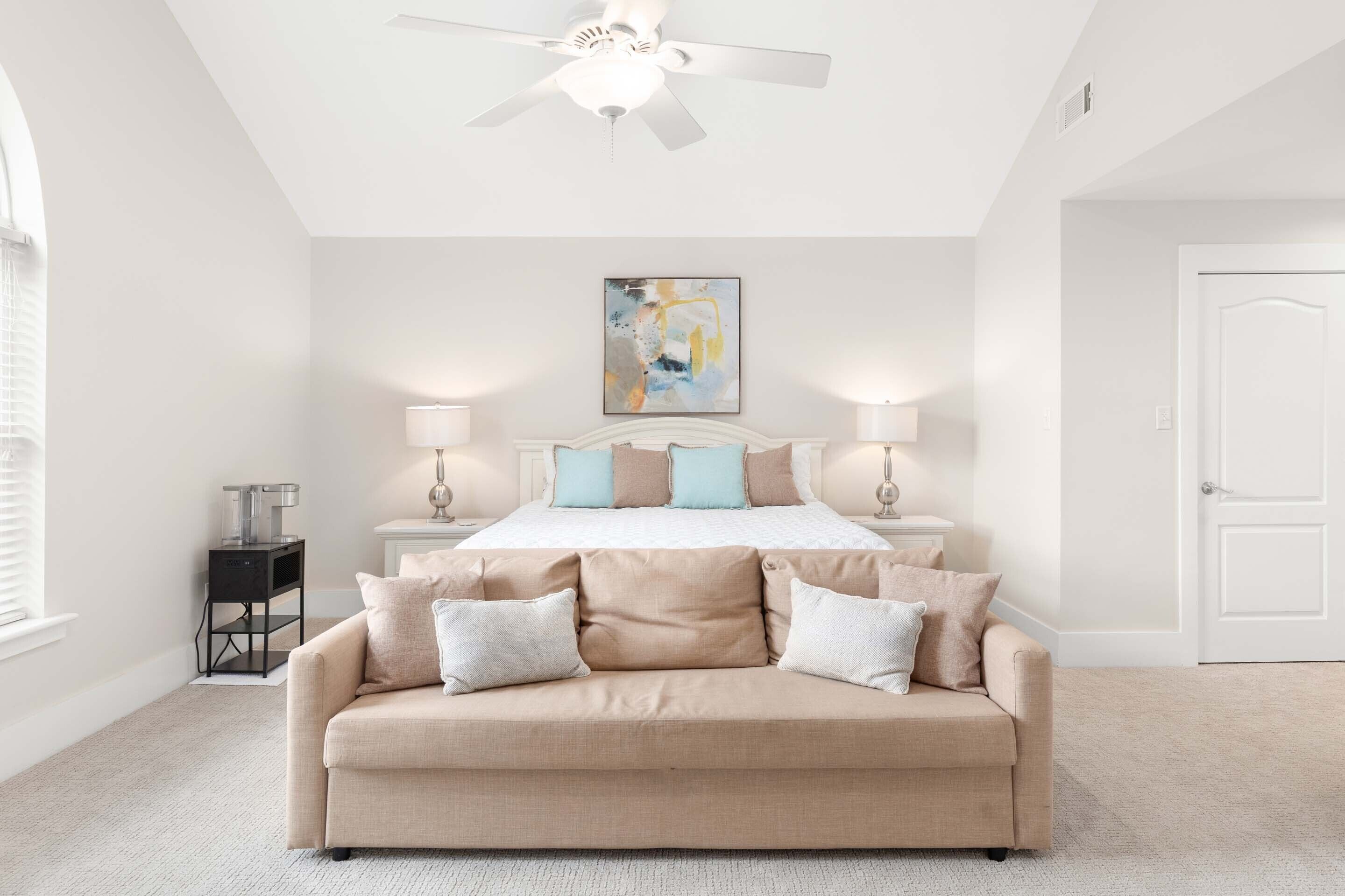 A bedroom with a bed, bedside tables, a ceiling fan, a painting, and a sofa.