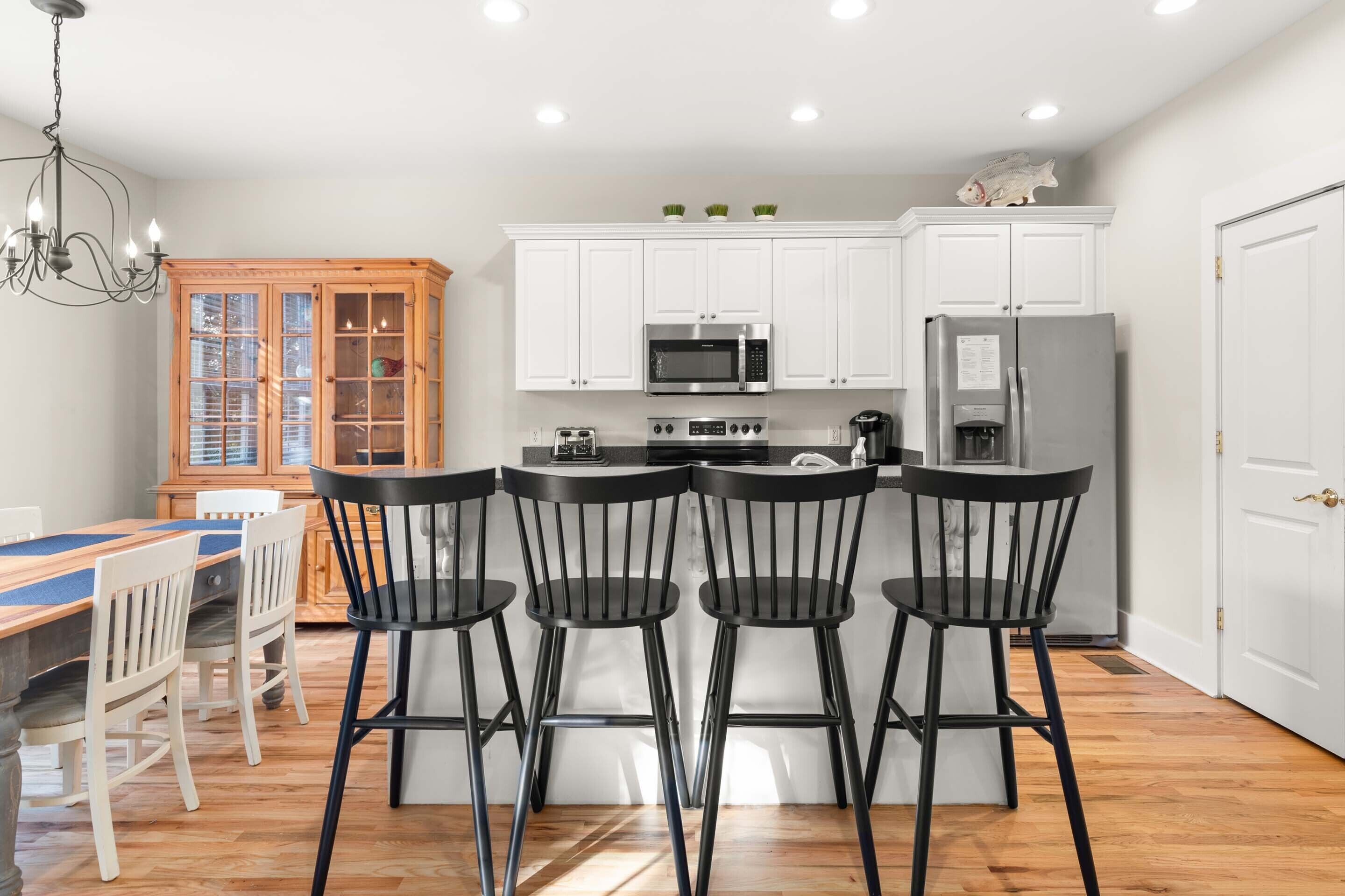 A modern kitchen with white cabinets, a stainless steel refrigerator, a microwave, and a stove. There are four black bar stools around the island.