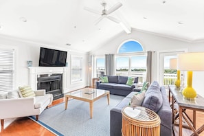 Relax in this spacious living room with a high ceiling, featuring a flat-screen TV above the fireplace, a large window with an outdoor view, and comfortable seating areas.