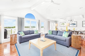 Relax in a spacious living area with a high ceiling and large windows offering a view of the outdoors. The room features two sofas, a coffee table, and a rug, as well as wicker side tables and a ceiling fan.