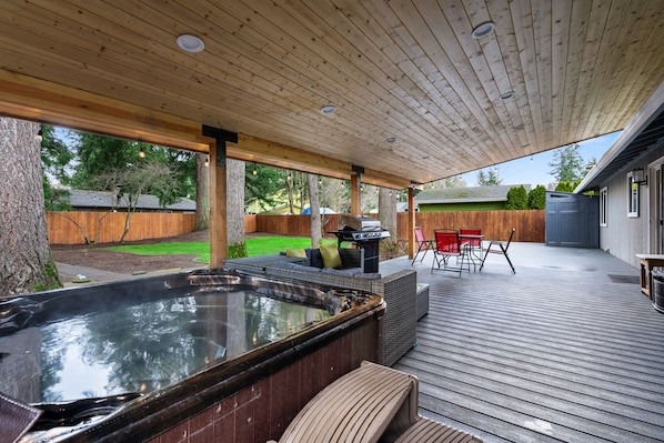 Un jacuzzi en una terraza de madera con un porche cubierto, mobiliario exterior y una parrilla.