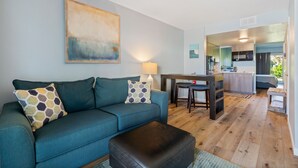 Un salon avec un canapé, une table basse et une lampe. On aperçoit un coin cuisine au fond de la pièce.