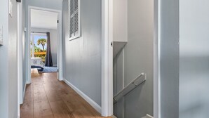 Un couloir doté d’un plancher en bois, un escalier avec des rampes en verre, et une vue sur un balcon avec une rambarde jaune et des palmiers.