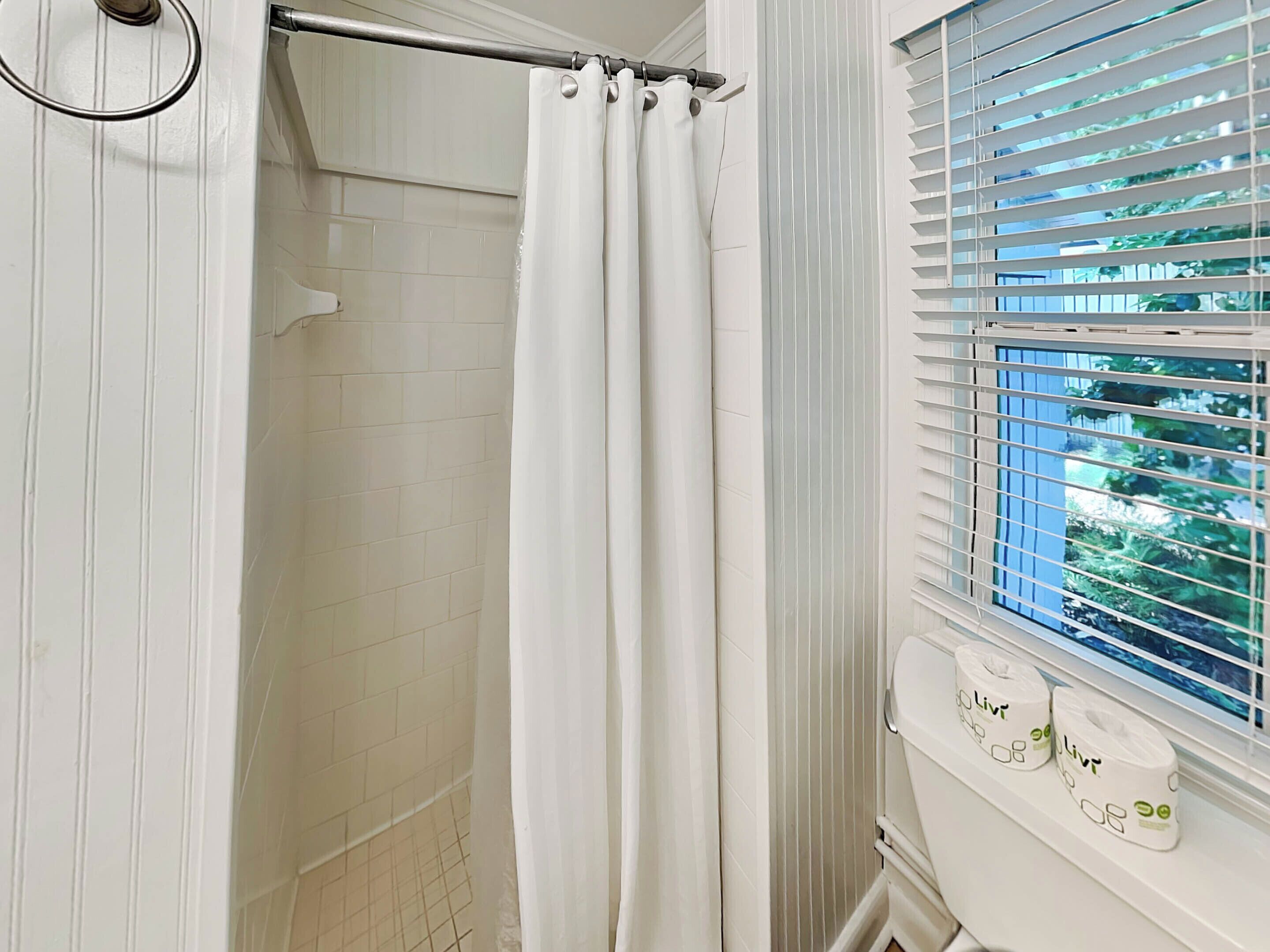 A bathroom with white tiled walls, a shower curtain, and a window with blinds.