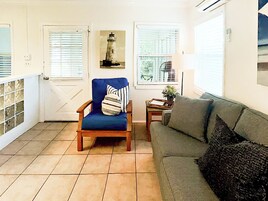 Une pièce comprenant une chaise en bois, un canapé gris, une petite table avec une plante et un tableau représentant un phare, accroché au mur.