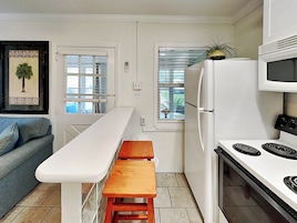 A compact kitchen with a white countertop, a wooden bench, a white refrigerator, a microwave, and a stove.