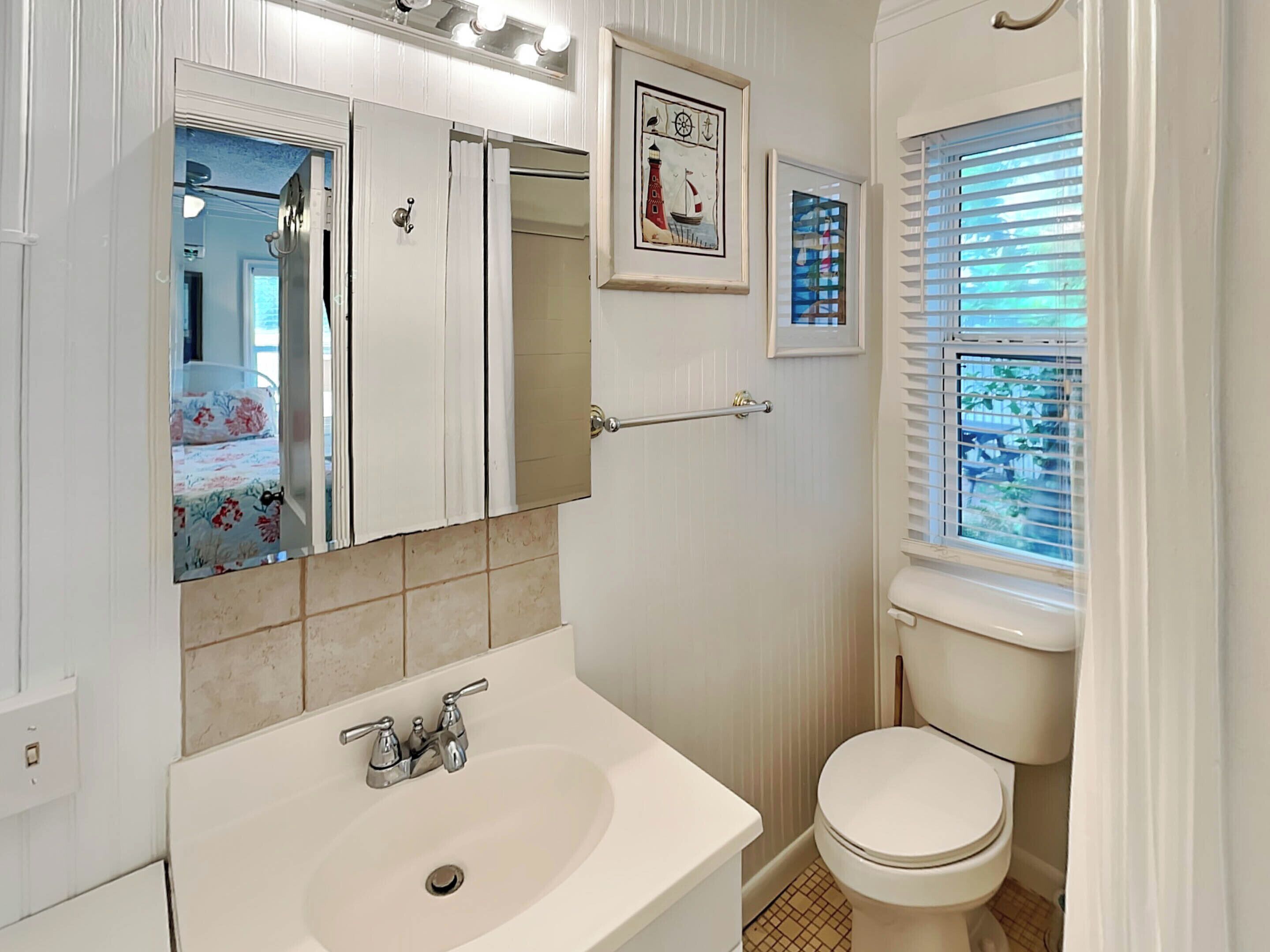 Ein Badezimmer mit einem weißen Waschbecken, einer Toilette und einem Spiegel. Es gibt ein Fenster mit Jalousien und ein gerahmtes Bild an der Wand.