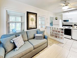 A living room with a grey sofa, a framed picture on the wall, and a kitchen area with white appliances.