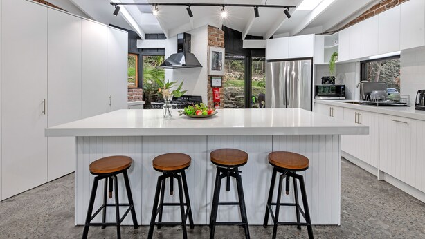 Una cocina moderna con una isla grande, tres taburetes, electrodomésticos de acero inoxidable y vistas al exterior.