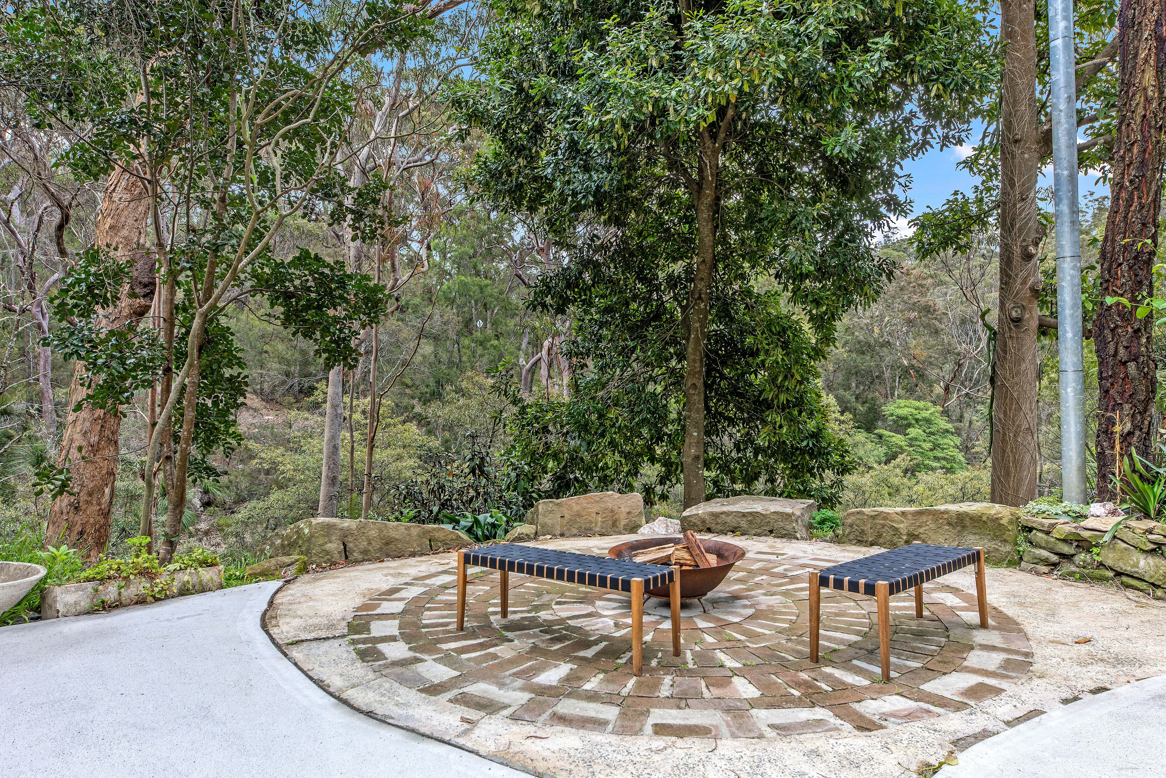 Un área de estar al aire libre con dos mesas de madera y bancos, rodeada por un patio circular empedrado de piedra, con árboles y rocas al fondo.