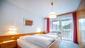 Une chambre d’hôtel comprenant un lit, une table de chevet, une lampe, une œuvre d’art, une fenêtre avec des rideaux et une vue sur la verdure.