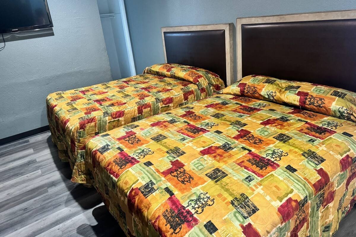 A room with two beds, each with a colorful quilt, a flat-screen TV mounted on the wall, and a wooden floor.