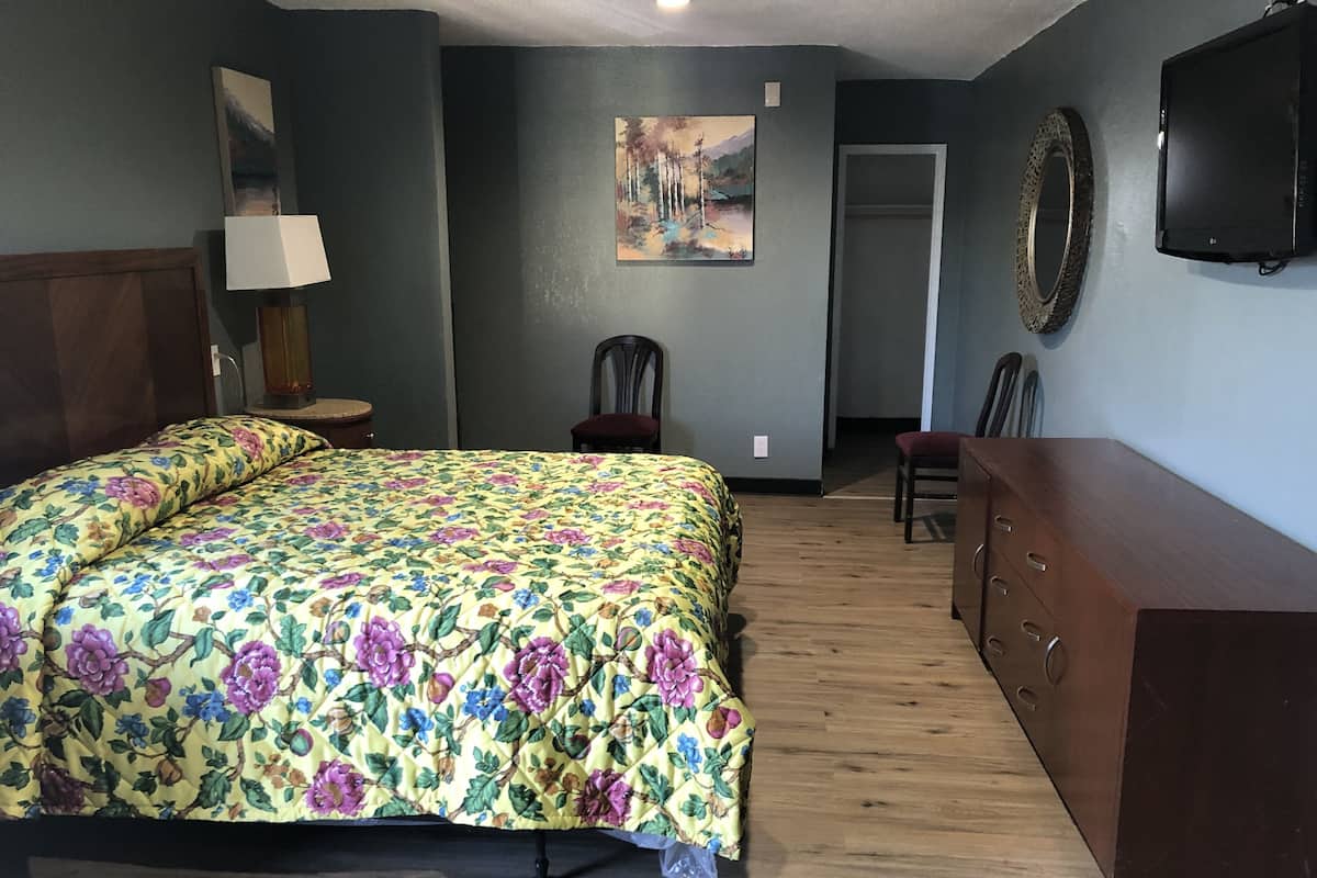 A bedroom with a floral bedspread, a wooden dresser, a chair, a desk, and a painting on the wall.