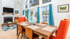 A spacious living room with a high ceiling, a fireplace, a television, and a dining area with a table set for four. - Catch A Wave by Tybee Vacation Rentals (Tybee Island)