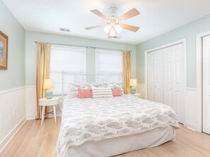 Une chambre à coucher comprenant un lit, deux tables de chevet, un ventilateur de plafond et une fenêtre avec des rideaux.