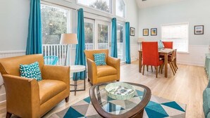 A spacious living room with large windows, a glass coffee table, and a dining area with chairs and a table. - Catch A Wave by Tybee Vacation Rentals (Tybee Island)