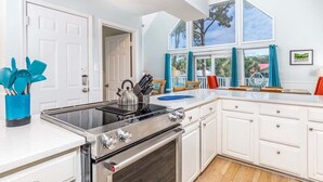 A modern kitchen with white cabinets, a stainless steel stove, and a large window with curtains. - Catch A Wave by Tybee Vacation Rentals (Tybee Island)