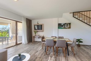 Un coin repas avec une table et des chaises en bois, un téléviseur fixé au mur et une vue sur l’extérieur grâce à une porte-fenêtre coulissante.