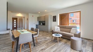 Uma sala de estar moderna com uma mesa de jantar de madeira, cadeiras estofadas cinzas, um sofá bege e uma pequena mesa redonda com uma planta.