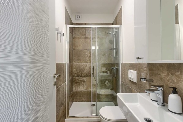A compact bathroom with a glass-enclosed shower, a toilet, and a sink with a soap dispenser.