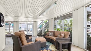 A covered outdoor seating area with wicker furniture, a patterned rug, and large windows offering views of the surrounding area. - Coastal Breezes by Tybee Vacation Rentals (Tybee Island)