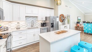 A modern kitchen with white cabinets, a central island, and a dining area with blue stools. - Coastal Breezes by Tybee Vacation Rentals (Tybee Island)
