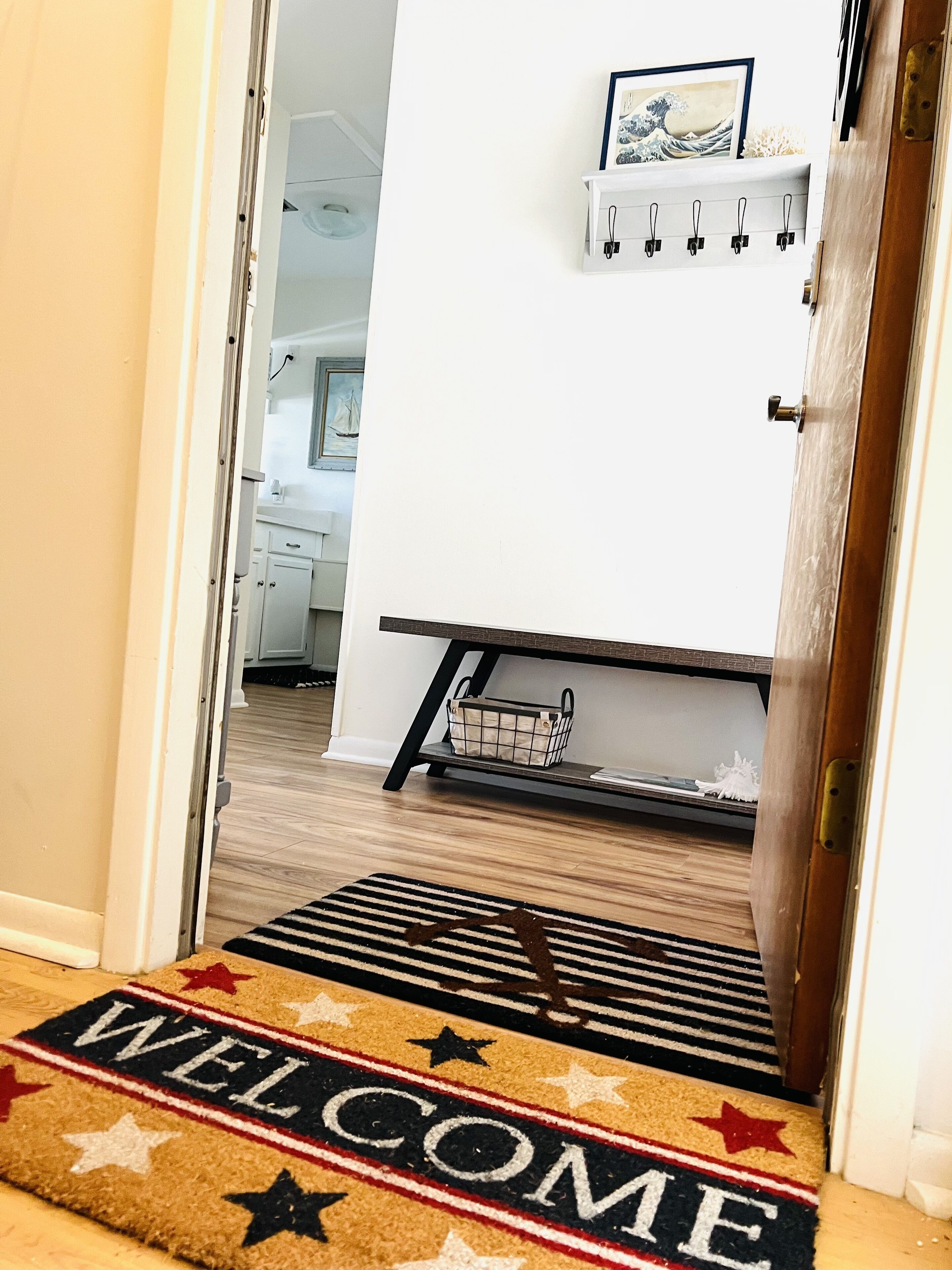 A welcome mat with 'WELCOME' and stars, a wooden floor, a kitchen area with cabinets, and a shelf with a framed picture.