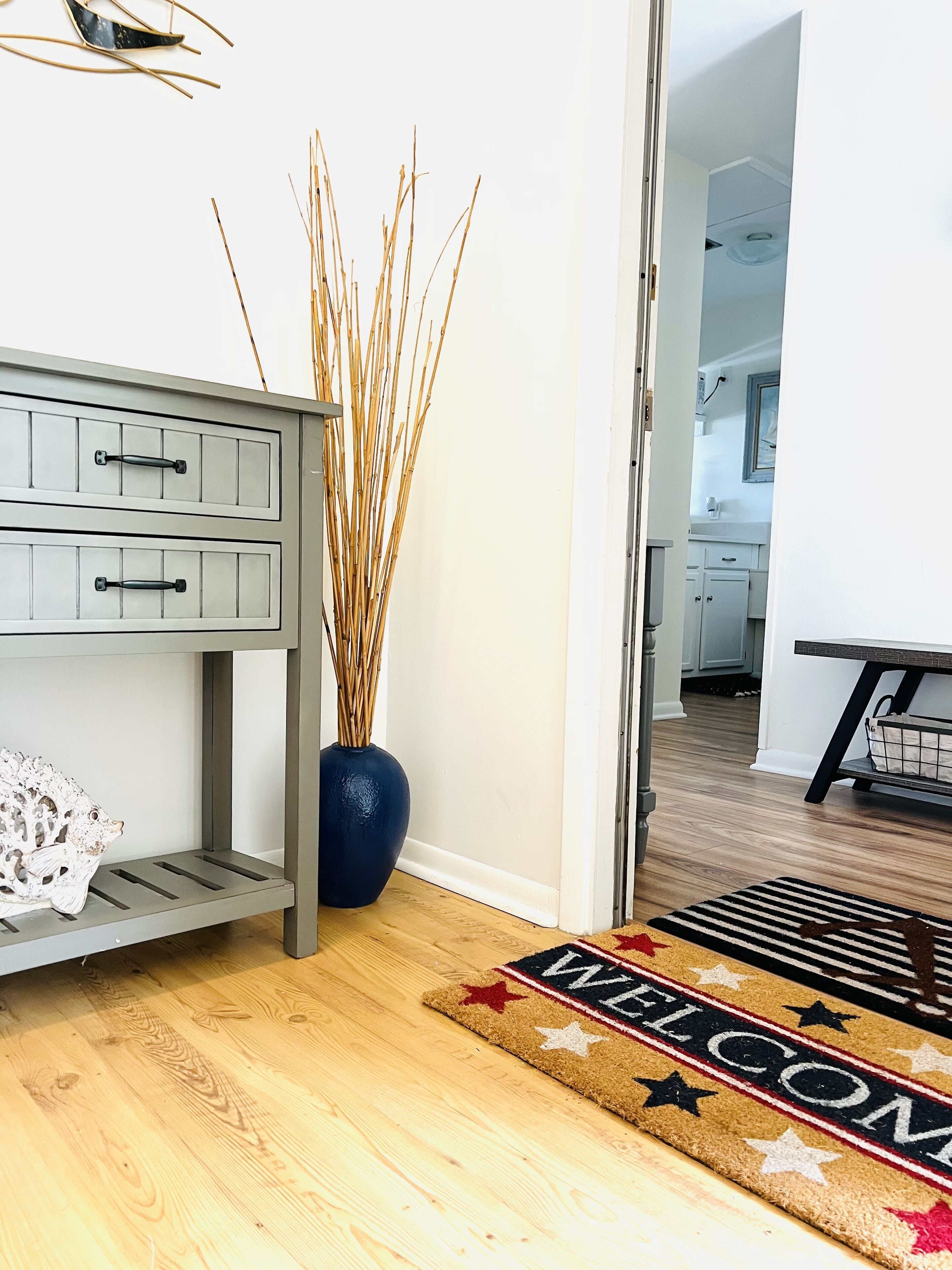 A wooden floor, a blue vase with branches, a gray console table, and a welcome mat.
