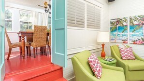 Una stanza luminosa con un tavolo e sedie in legno, una poltrona verde, una scala rossa e un quadro raffigurante una scena tropicale.