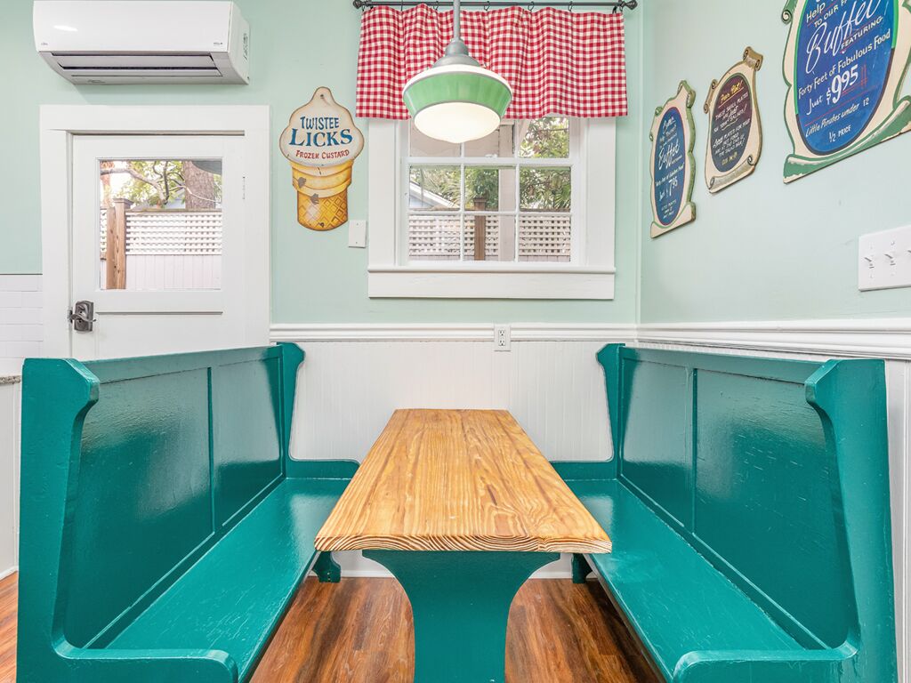Una habitación con bancos pintados de verde azulado y una mesa de madera, una ventana con cortina a cuadros y adornos en la pared, incluyendo un cartel de Twistee Licks y dos menús enmarcados.