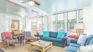 A spacious living room with a blue sofa, striped chairs, a coffee table, and large windows offering a view of trees. - Coquina Cottage by Tybee Vacation Rentals (Tybee Island)