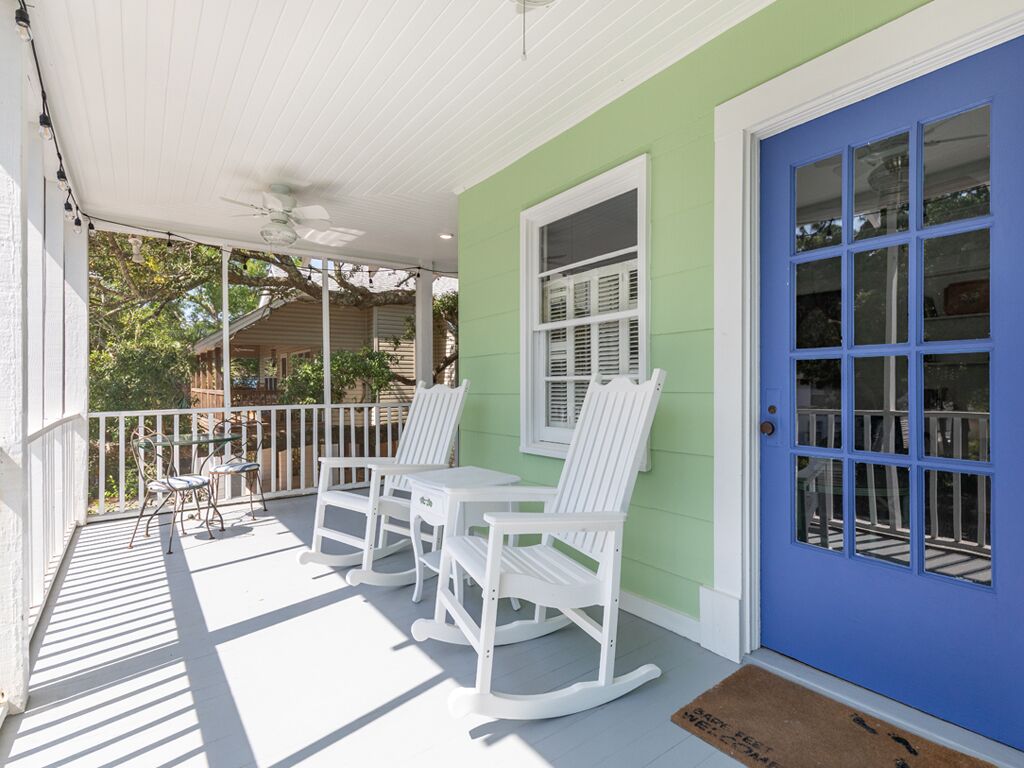 A porch with white rocking chairs and tables, a blue door, and a white ceiling fan.