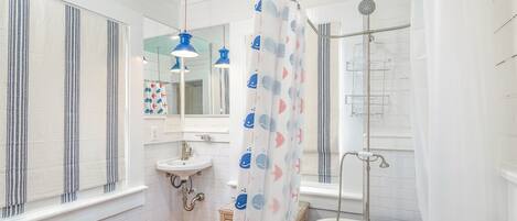 A bathroom with a white bathtub, a shower, a sink, and a window with striped curtains.