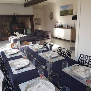 A dining area with a long table set for a meal, a fireplace, a sofa, and a kitchen area in the background.