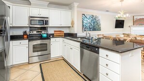 A modern kitchen with white cabinets, stainless steel appliances, a large island, and a dining area with wooden chairs. - Dolphin Watch 6 by Tybee Vacation Rentals (Tybee Island)
