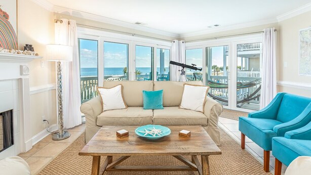 A spacious living room with a large window overlooking a balcony, a wooden coffee table with decorative items, and a teal armchair. - Dolphin Watch 6 by Tybee Vacation Rentals (Tybee Island)