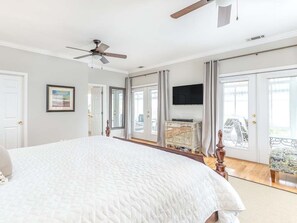 A bedroom with a large bed, a ceiling fan, a TV, and a balcony with curtains.