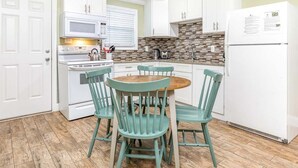 A compact kitchen with white cabinets, a microwave, a stove, a refrigerator, and a wooden dining table with four chairs. - Dune A by Tybee Vacation Rentals (Tybee Island)
