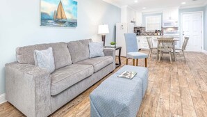 A living room with a grey sofa, a blue ottoman, and a painting of a sailboat. - Dune B by Tybee Vacation Rentals (Tybee Island)
