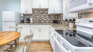 A modern kitchen with white cabinets, a granite countertop, a built-in stove, and a dining area with a wooden table and chairs. - Dune B by Tybee Vacation Rentals (Tybee Island)