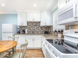Una cocina moderna con armarios blancos, encimera de granito, estufa integrada y un comedor con mesa y sillas de madera.