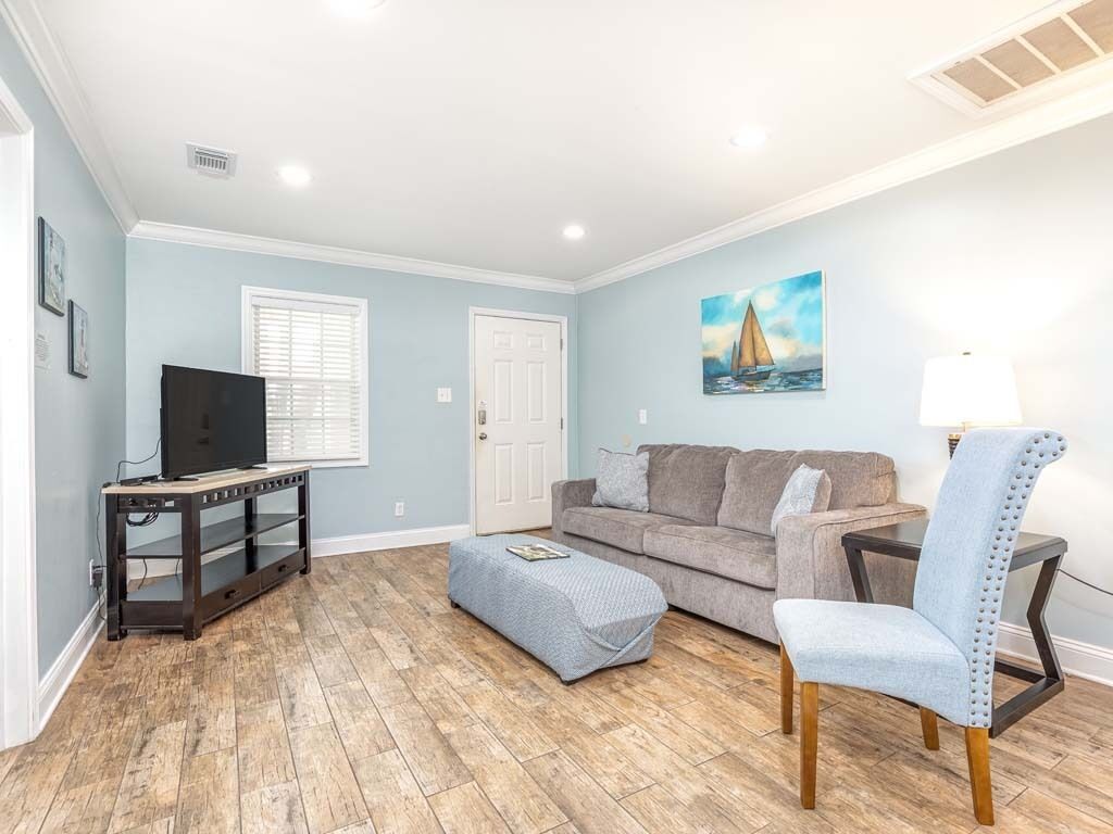 A living room with a grey sofa, a blue ottoman, a TV on a wooden stand, and a painting on the wall.