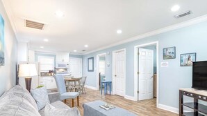 A living room with a grey sofa, a coffee table, and a TV stand. There is a kitchen in the background with white cabinets and a dining area with a table and chairs. - Dune B by Tybee Vacation Rentals (Tybee Island)